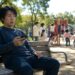 Man on a park bench looking at his phone with a playground in the background.